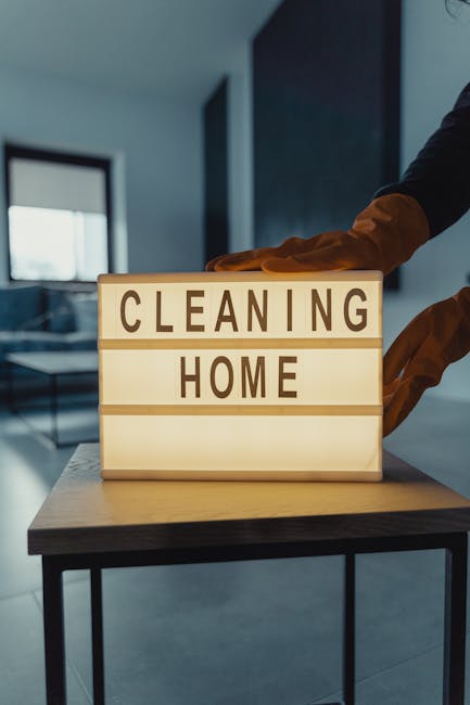 A person wearing brown cleaning gloves is placing a lightbox sign with black letters reading 'CLEANING HOME' on a table or countertop, indicating a focus on domestic cleaning services. The background 