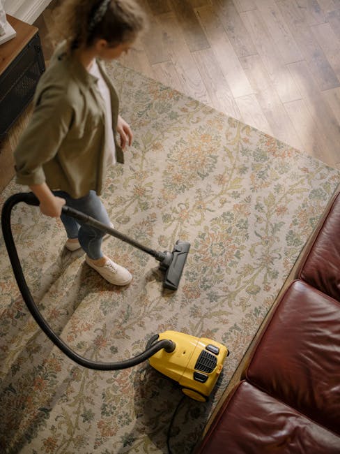 A woman using a yellow vacuum cleaner on a patterned, beige-and-olive carpet in a living room. She is dressed in casual clothing, including a beige jacket, jeans, and white sneakers. The room features hardwood flooring adjacent to the carpet, a red leather sofa visible at the edge of the image, and soft, natural lighting. The scene depicts surface cleaning and deep cleaning in a domestic setting, highlighting the professional cleaning services provided by Cleaner Enfield as part of their carpet cleaning service in Palmers Green, Enfield.