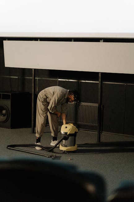 A person kneeling on a patterned carpet in a room with warm lighting, using a yellow and black vacuum cleaner for surface cleaning. They are placing or retrieving a cleaning cloth or accessory from the vacuum's open lid. The room features a wooden surface or furniture in the background, and the carpet appears clean and well-maintained. This scene illustrates domestic cleaning activities associated with professional and deep cleaning services provided by Cleaner Enfield, targeting hygiene and maintenance for residential spaces.