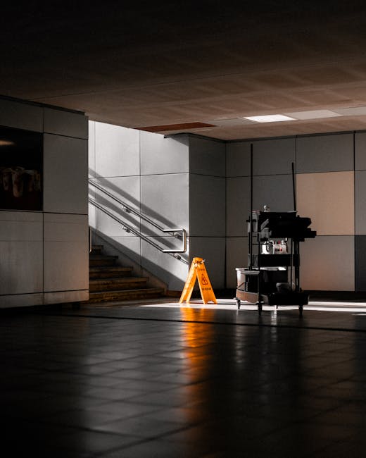 A room with a newly finished wooden floor during a deep cleaning process, with a commercial vacuum cleaner placed on the floor and a broom lying nearby. The background shows a white wall with some con